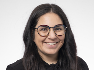 Woman with long dark hair, wearing large round glasses and a black top, smiling at the camera against a plain white background.