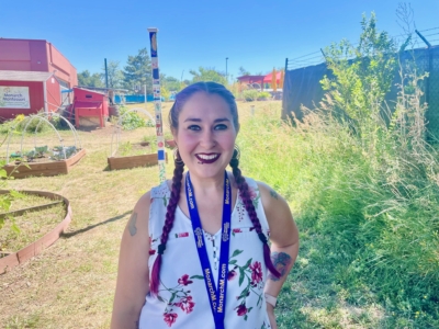 A woman with braided hair, wearing a white floral top and a blue lanyard, smiles outside in a sunny garden area with greenery and raised garden beds in the background.