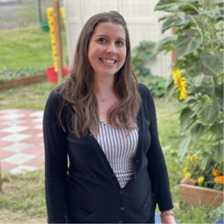 A woman with long brown hair in a black cardigan and striped top smiles while standing outdoors near sunflowers and greenery.