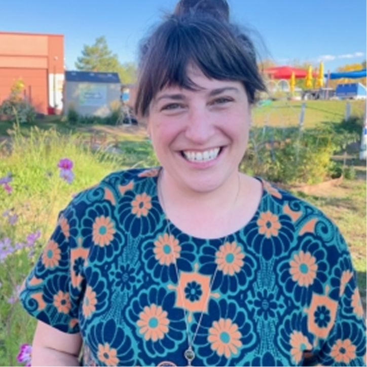 A woman with dark hair in a bun smiles outdoors, wearing a floral-patterned top. She stands in front of greenery, flowers, and buildings under a clear blue sky.