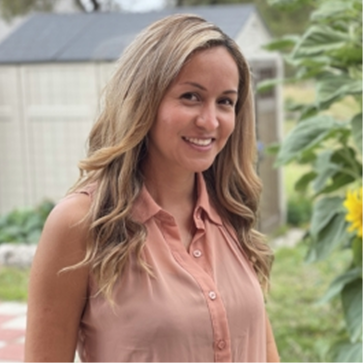 Woman with long, wavy blonde hair wearing a sleeveless peach blouse, smiling outdoors near a sunflower and garden shed.